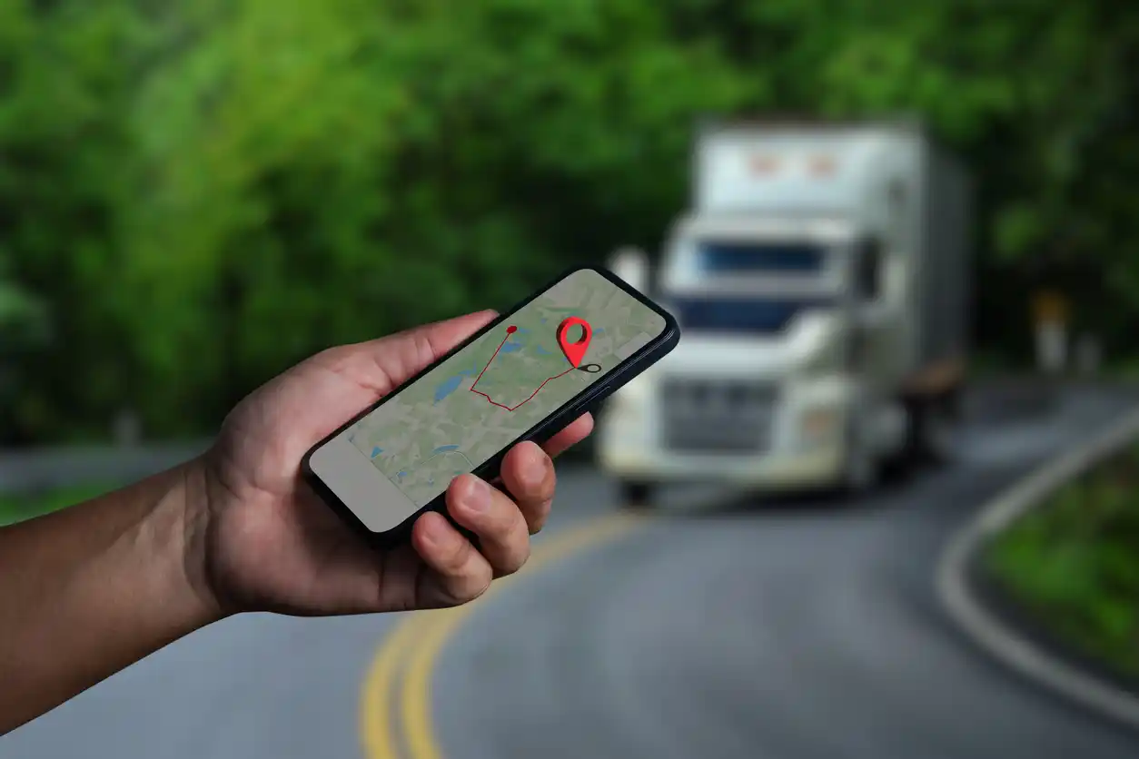 Hand holding a smartphone with a GPS map pin while a semi truck drives a winding road, tracking a last-mile delivery
